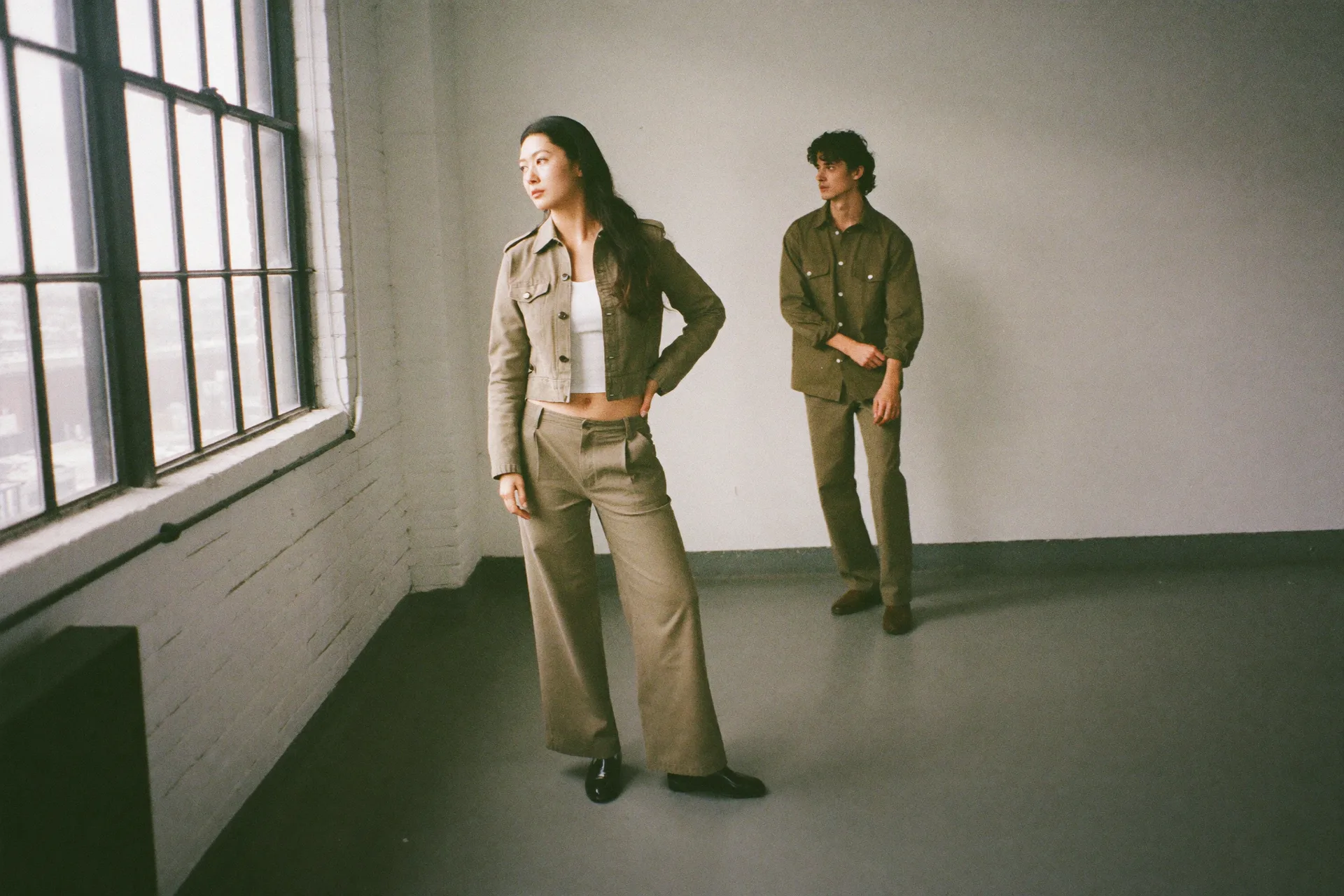 A woman and a man in a daylight loft in two cuts of the same olive look: she stands by the windows in a cropped utility jacket, white tank and wide-leg pants; he waits a step behind in a loose overshirt and matching pants, fastening a cuff.