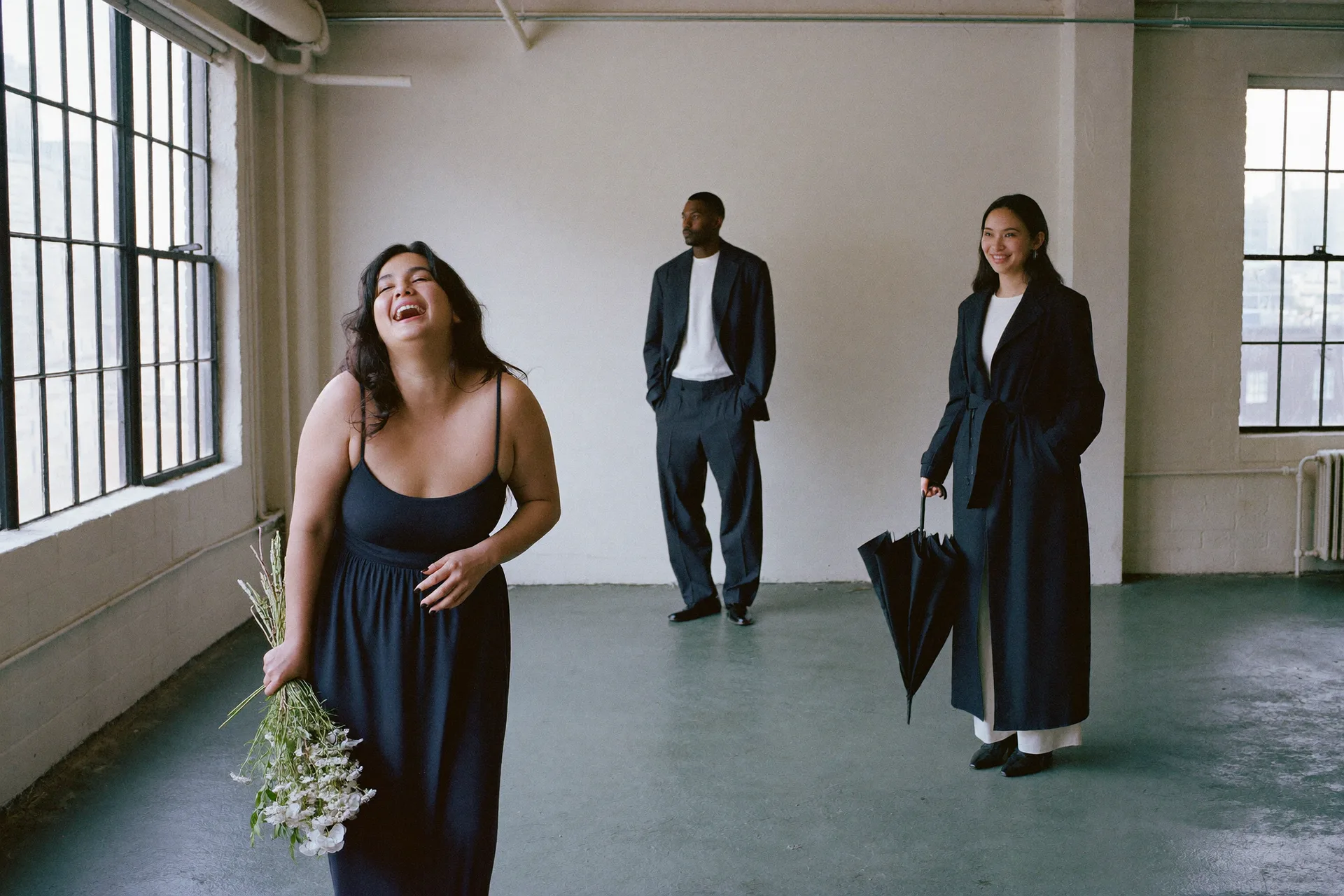 Two women and a man stand apart in a daylight loft, each dressed for a different occasion in the same deep navy: a slip dress with a bunch of white flowers, a relaxed trouser suit, and a long belted trench with a furled umbrella.