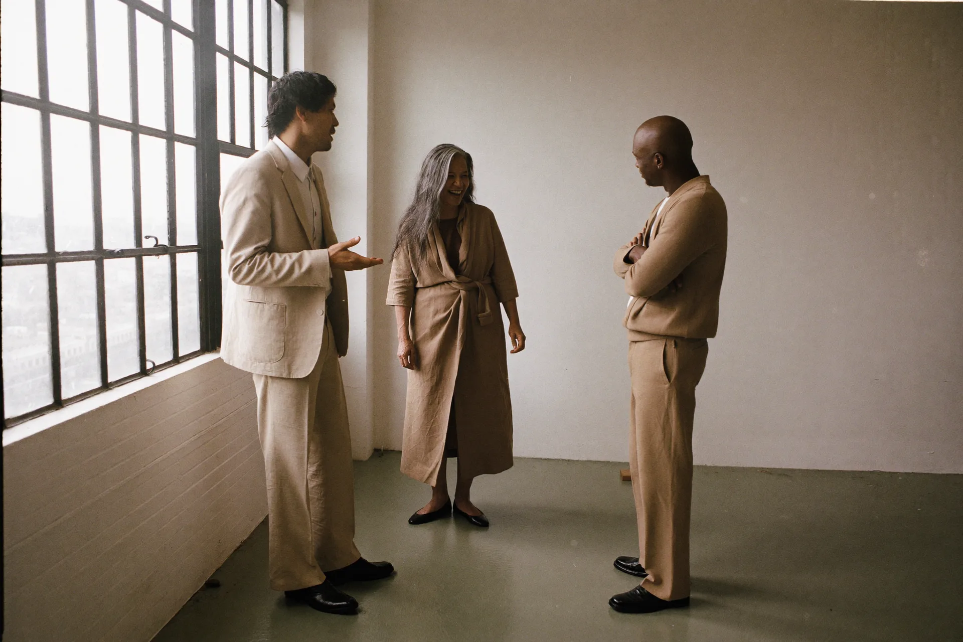 Three people mid-conversation by the loft windows, all in driftwood linen: a young man in a suit talking with an open hand, a gray-haired woman in a wrap dress laughing, and an older man in a cardigan listening with his arms crossed.