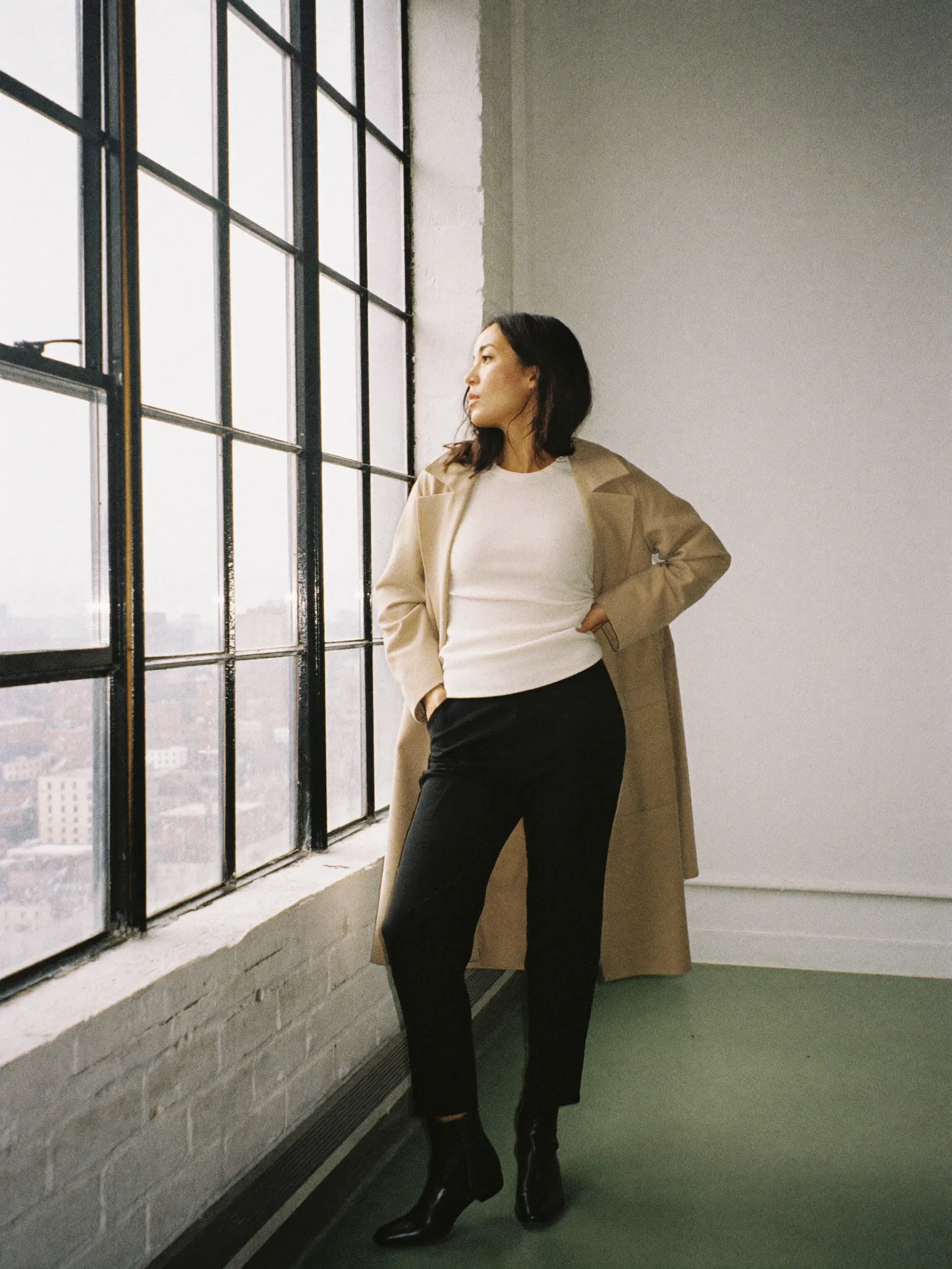 A woman in a sand-colored coat over an ivory knit stands by the loft windows, turned toward the daylight.
