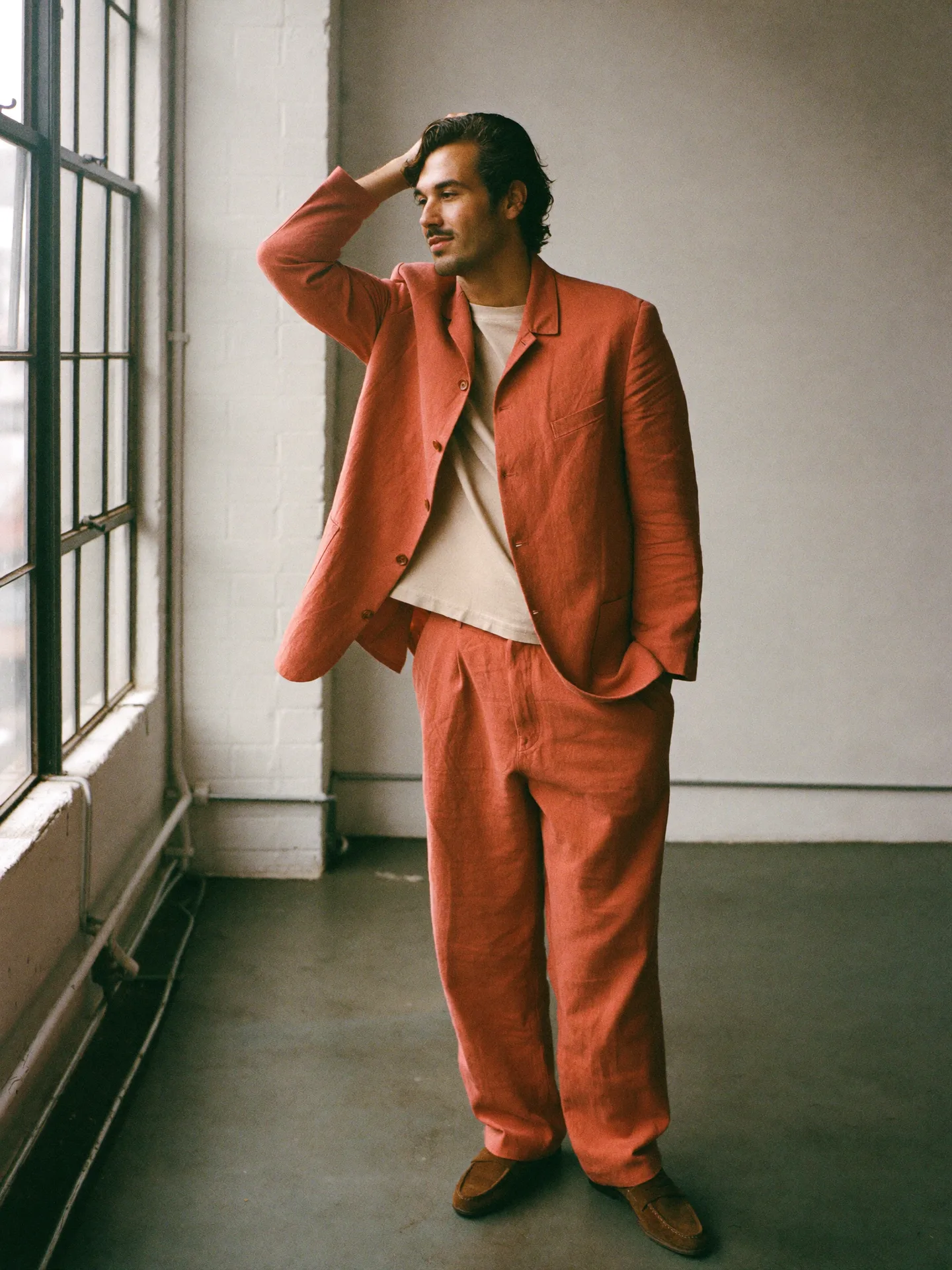 A man with golden olive skin in a tomato-coral linen suit over an ecru T-shirt, glancing toward the loft windows.